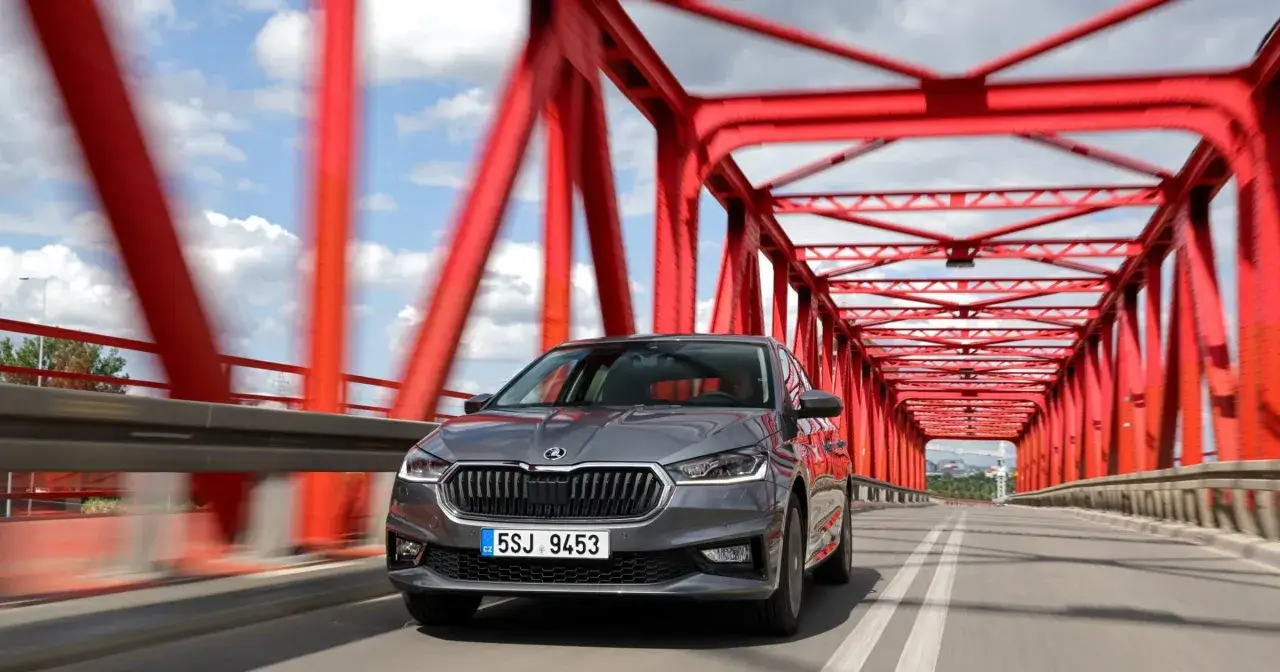 Szara Skoda Fabia 1 jedzie przez czerwony most, chmury na niebie tworzą dynamiczne tło.