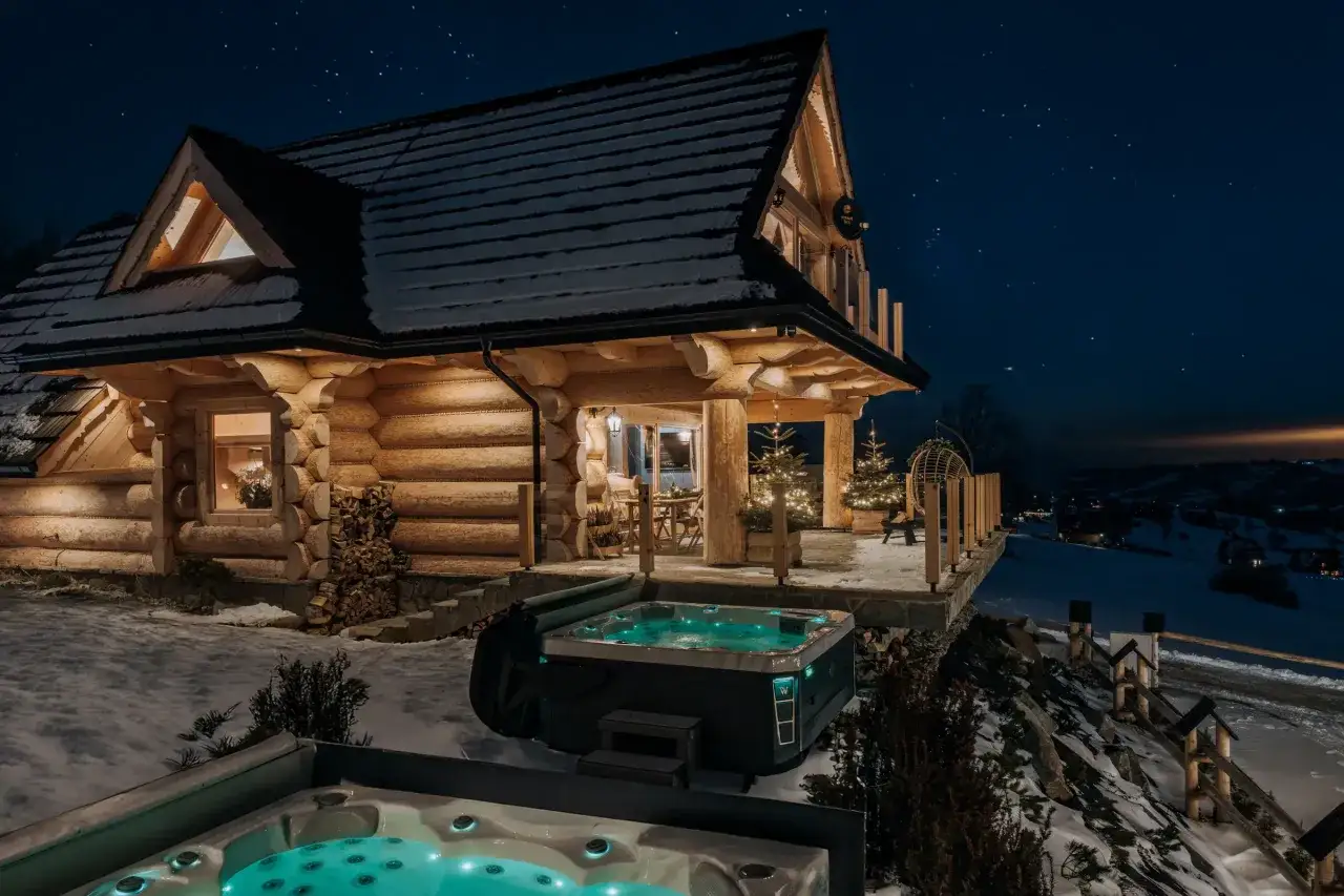 Drewniany bukowina tatrzańska domek nocą, oświetlony, ze śniegiem na dachu i dwoma jacuzzi. Gwiaździste niebo nad Tatrami.