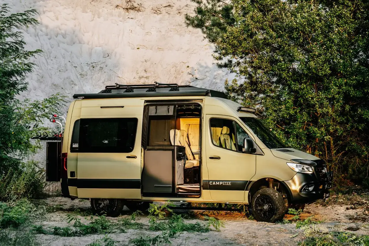 Beżowe kampery terenowe Campster gotowe na przygodę. Otwarty pojazd z widocznym wnętrzem, otoczony piaskiem i zielenią.