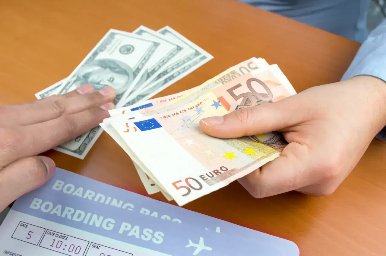 Wypłata z bankomatu za granicą: ręka trzyma euro, obok leżą dolary i karta pokładowa.