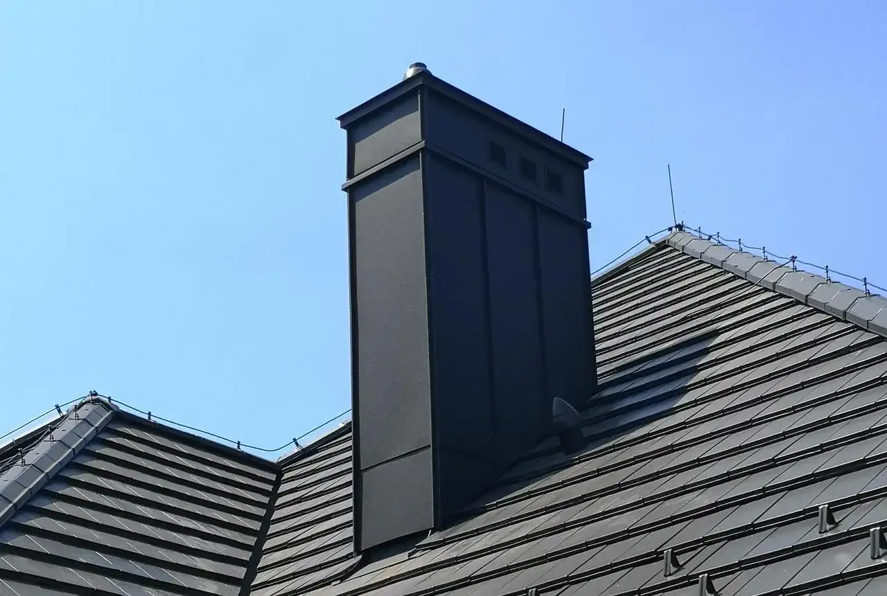 Nowoczesny, czarny komin g&oacute;ruje nad szarym dachem. Wok&oacute;ł komina biegnie instalacja odgromowa, chroniąc budynek przed burzą.