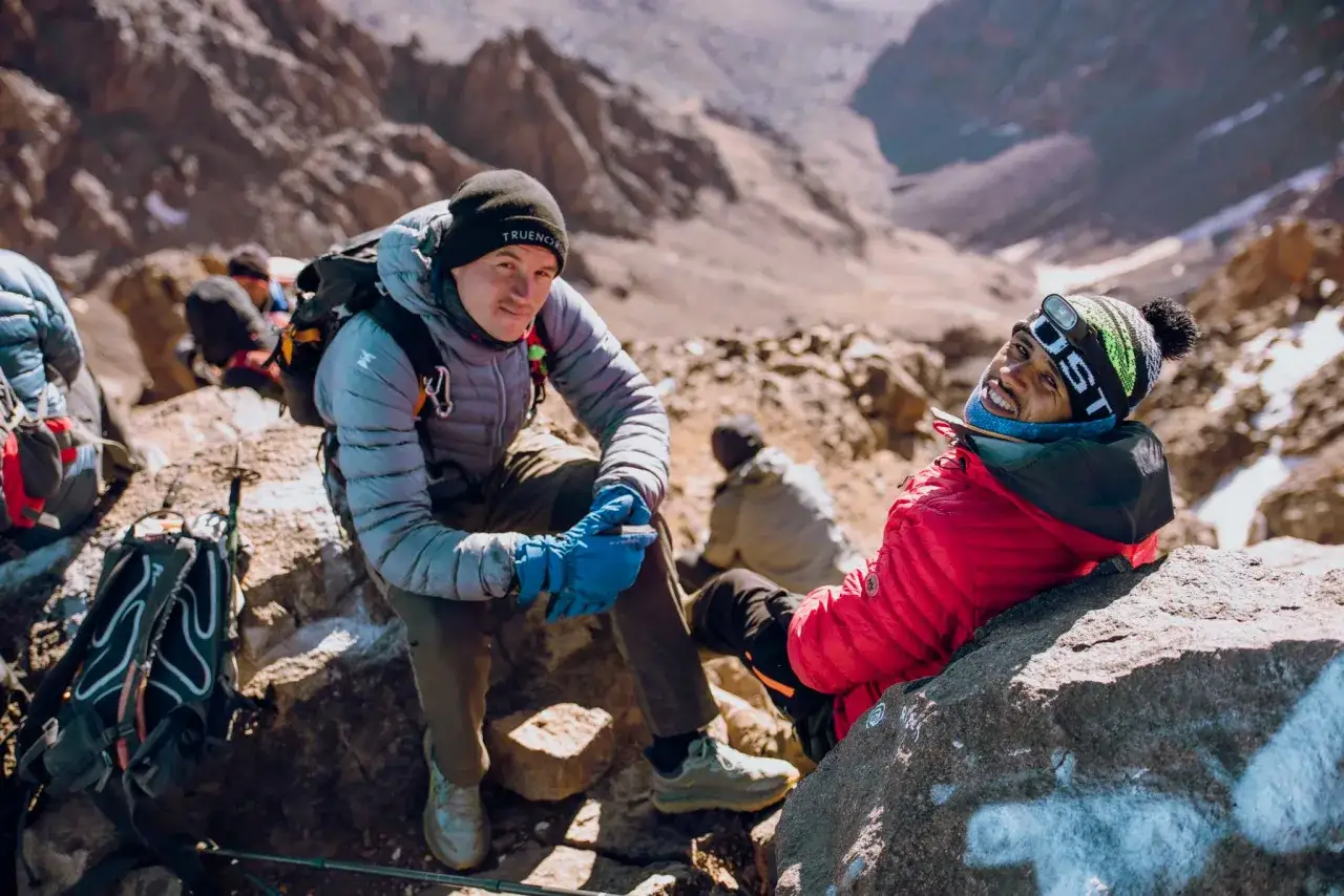 Uśmiechnięci alpiniści na Toubkal wyprawa, odpoczywają na kamienistym zboczu.