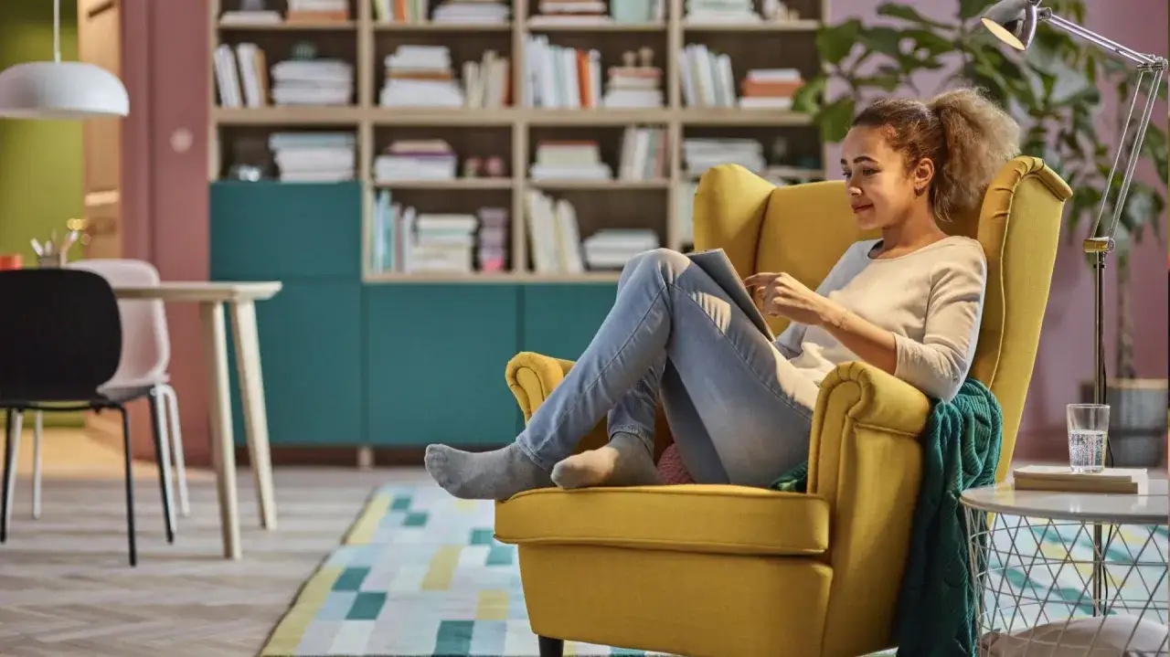 Kobieta relaksuje się z tabletem w ż&oacute;łtym fotelu, tworząc przytulny kącik do czytania. W tle regały z książkami i kolorowy dywan.