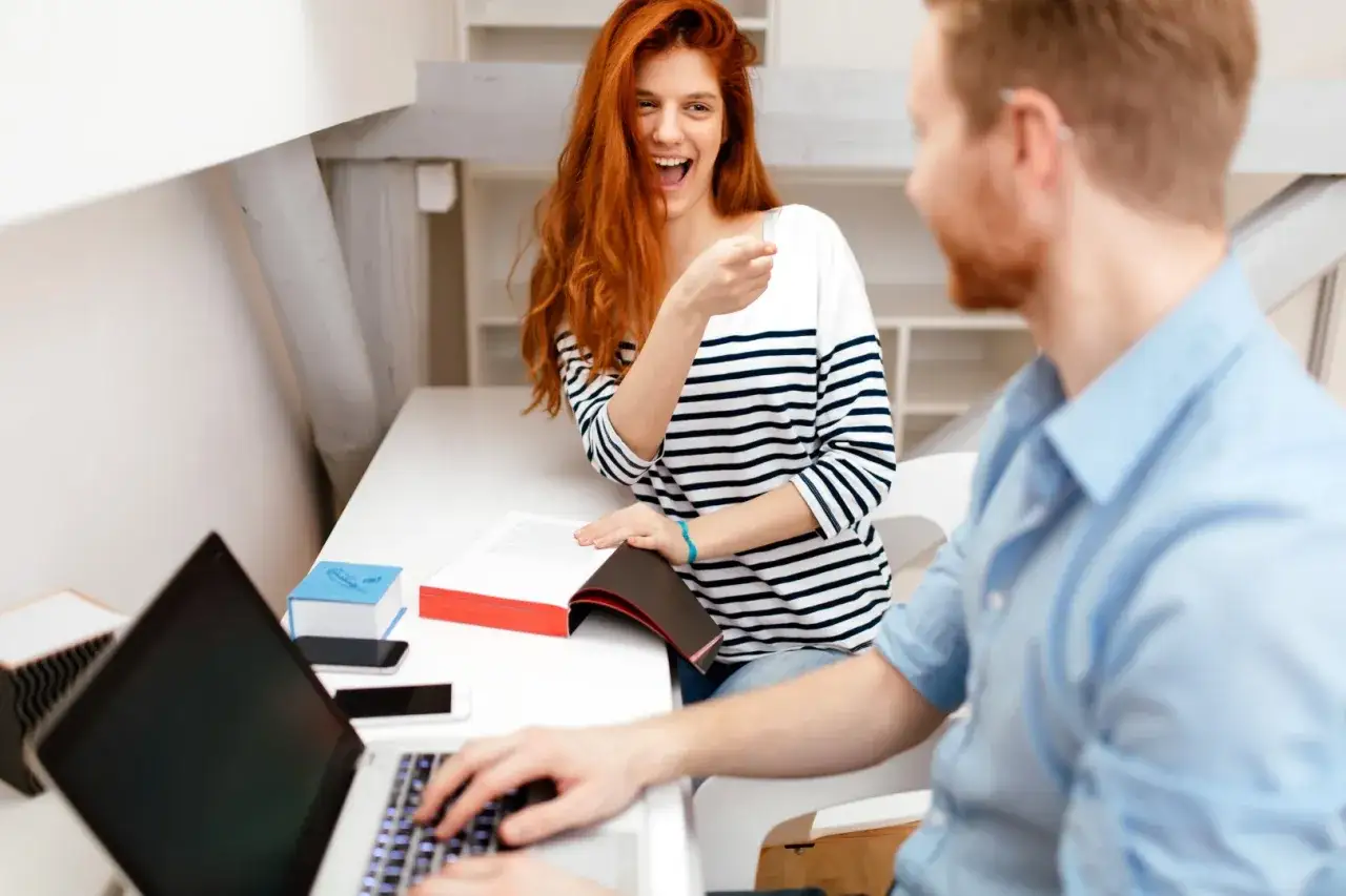 Kobieta z rudymi włosami śmieje się i wskazuje palcem, gdy mężczyzna pracuje przy laptopie. Czy to objawy zakochanego szefa?
