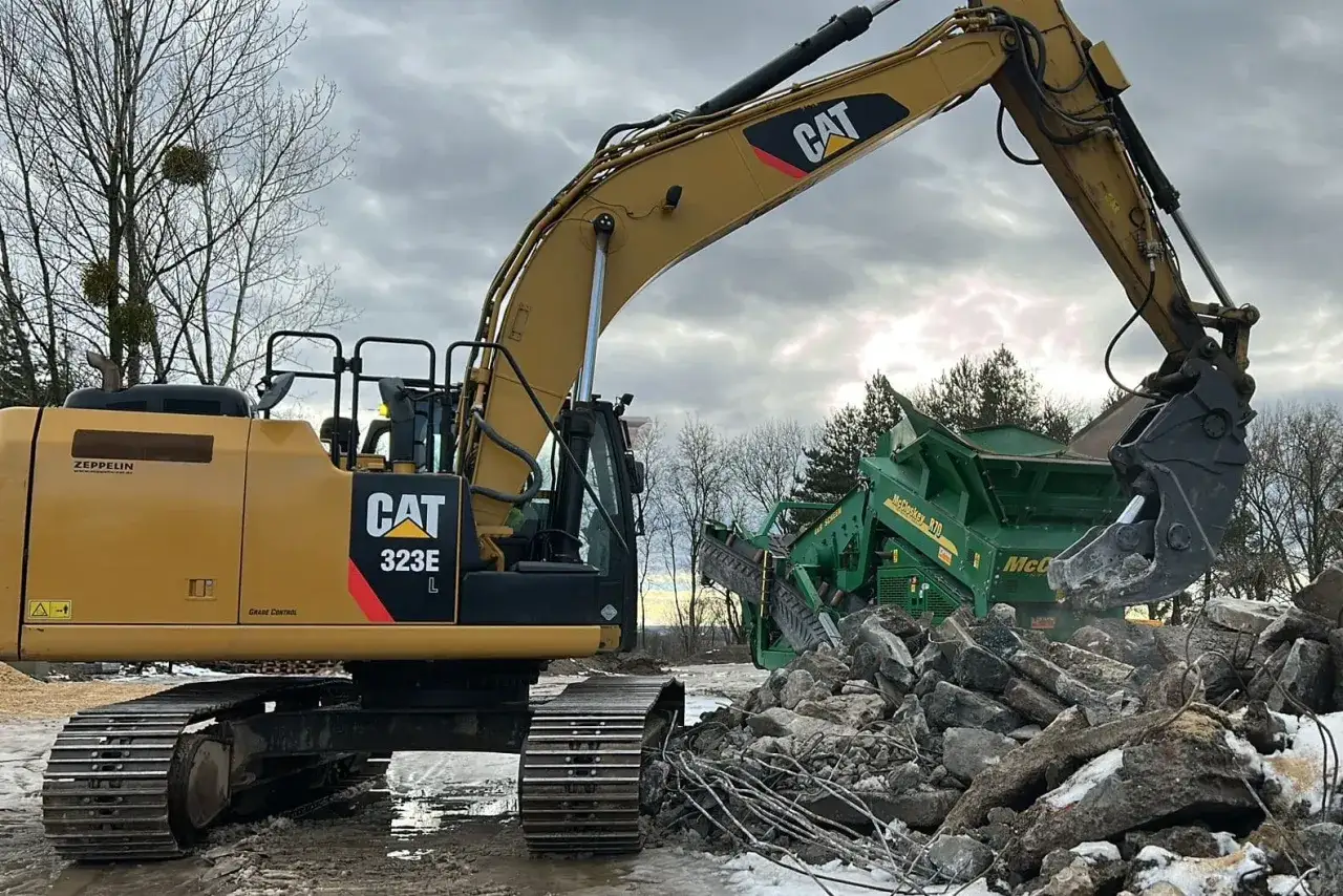 Żółta koparka CAT 323E pracuje przy kruszarce. Zastanawiasz się, ile kosztuje koparka? To potężne maszyny budowlane.