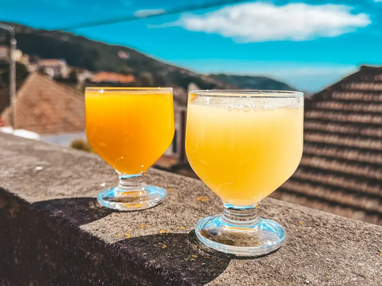 Dwa kieliszki z orzeźwiającym napojem, być może z dodatkiem rumu z Madery, na tle błękitnego nieba i zielonych wzgórz.