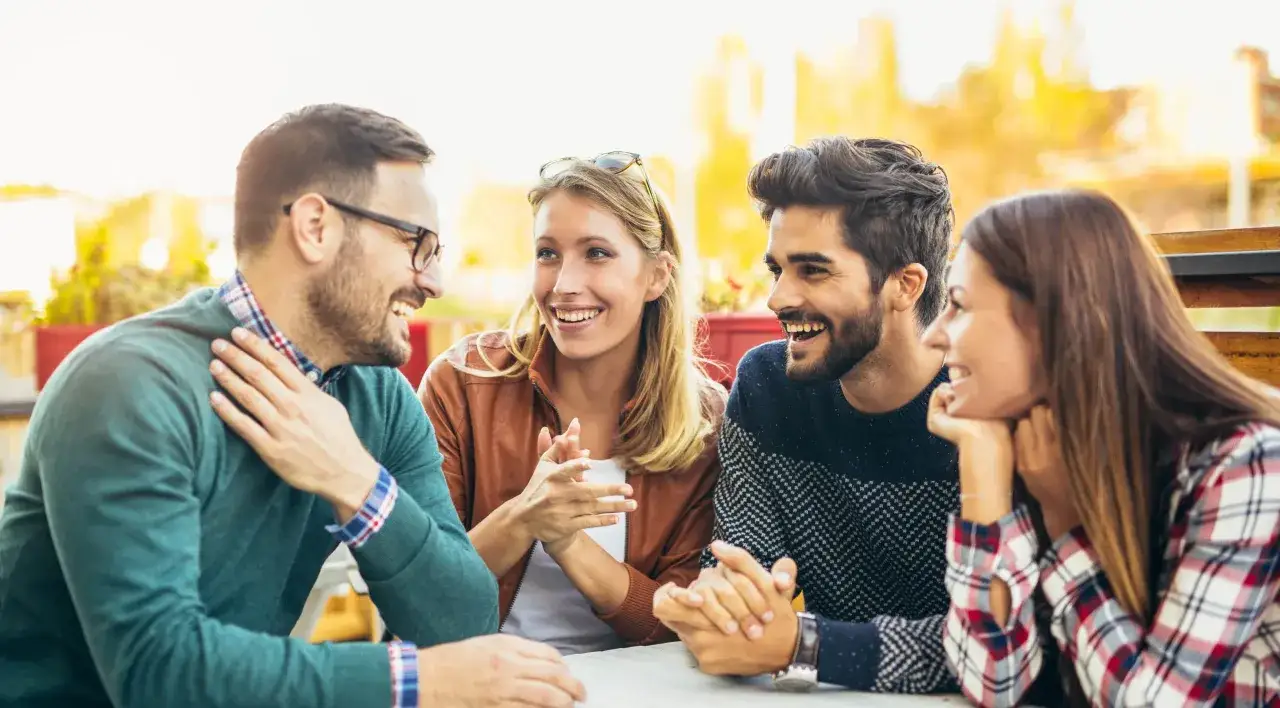 Dw&oacute;ch mężczyzn i dwie kobiety śmieją się przy stole. R&oacute;żne typy mężczyzn, jeden w okularach, drugi z brodą, rozmawiają z kobietami.