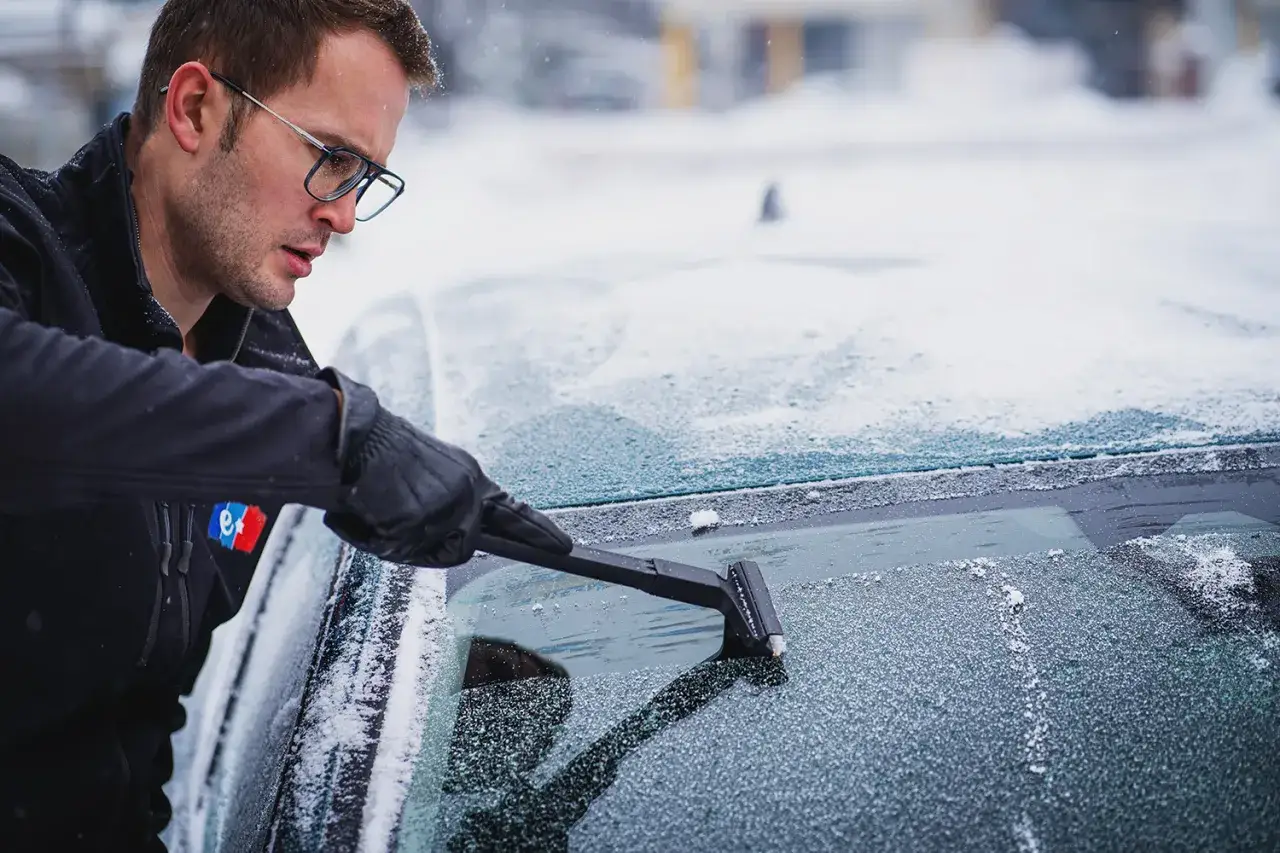 Mężczyzna w okularach i rękawiczkach z determinacją usuwa l&oacute;d z przedniej szyby auta, pokazując, jak odmrozić szyby w aucie.