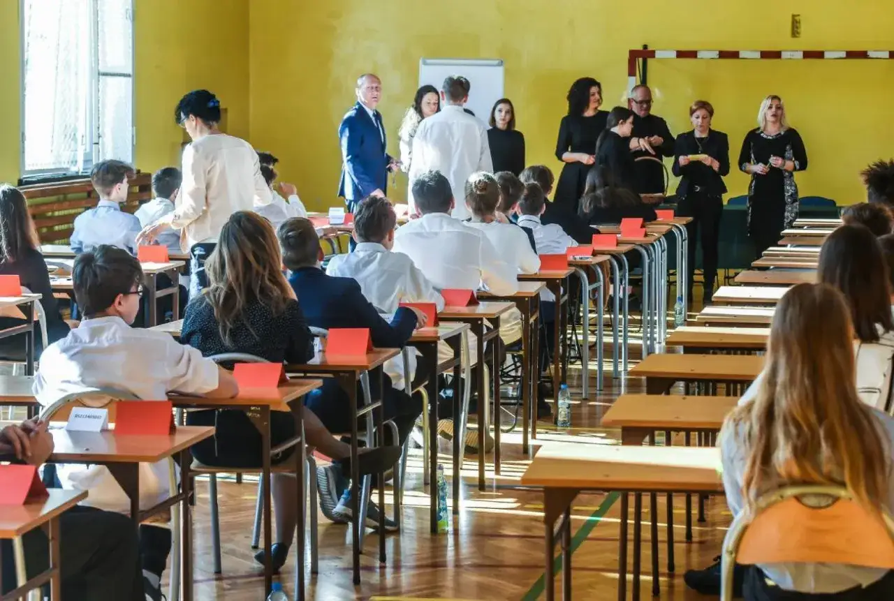 Uczniowie ósmej klasy siedzą w ławkach, gotowi do egzaminu. Nauczyciele obserwują salę.