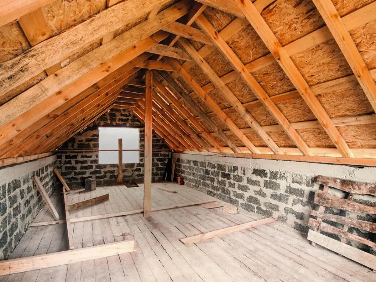 Widok wnętrza poddasza w trakcie budowy. Drewniany stelaż dachu i ściany z bloczk&oacute;w betonowych. Widać deski podłogowe i materiały budowlane. To początek prac, jak zrobić sufit na poddaszu.