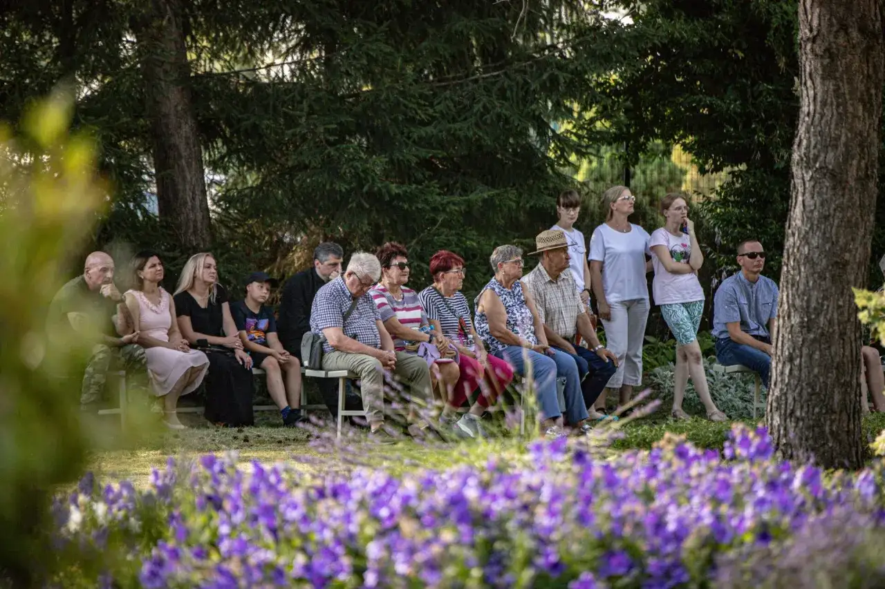 Ludzie siedzą na ławkach w ogrodzie, słuchając czegoś. Pamiętajmy o ogrodach, bo to piękne miejsca spotkań.