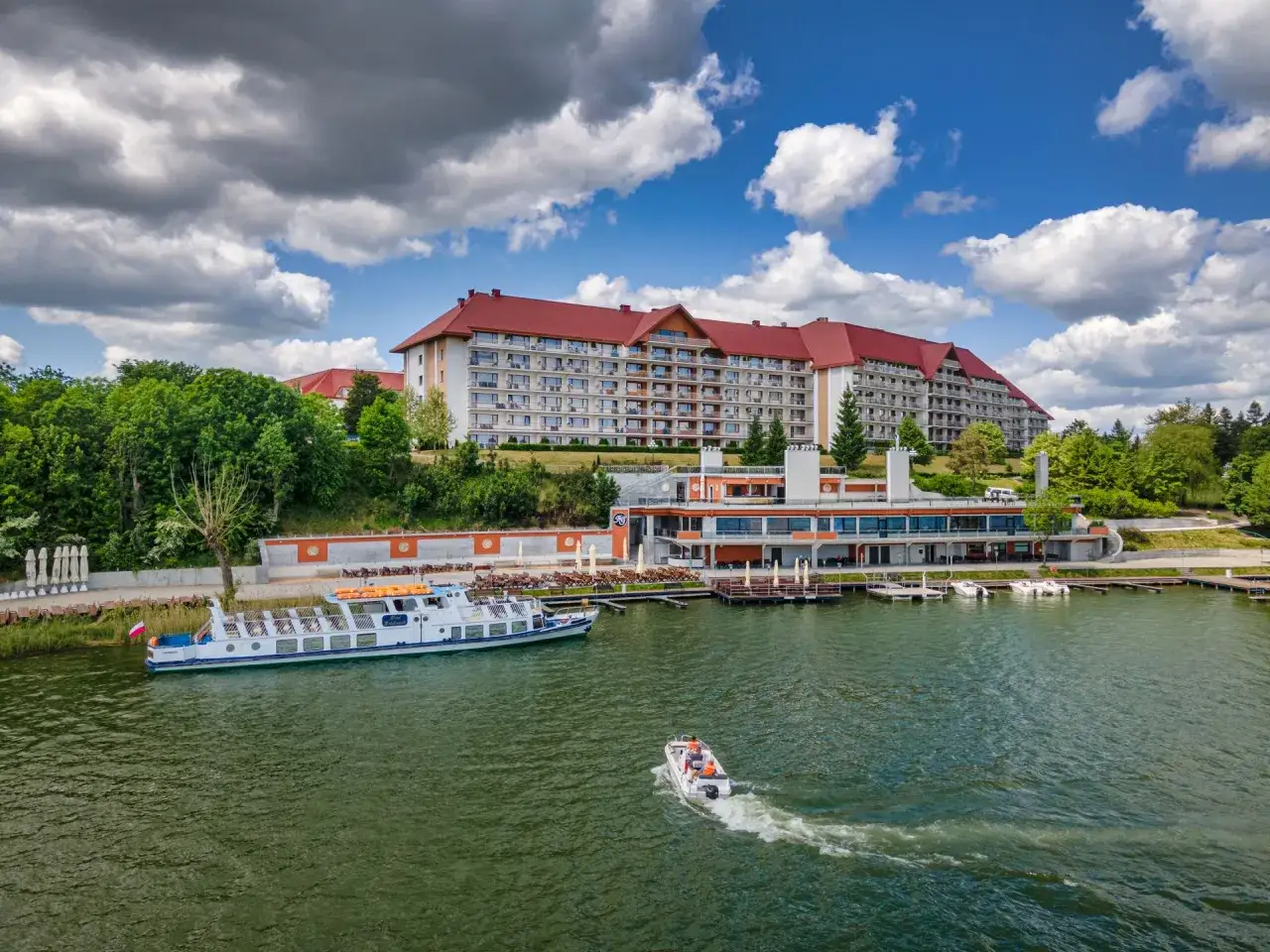 Wielki kompleks hotelowy Gołębiewski nad jeziorem, z białym statkiem wycieczkowym i motorówką na wodzie.