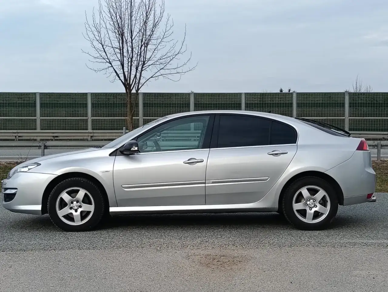 Srebrne Renault Laguna 3 stoi na tle ekranów akustycznych i nagiej drzewa.