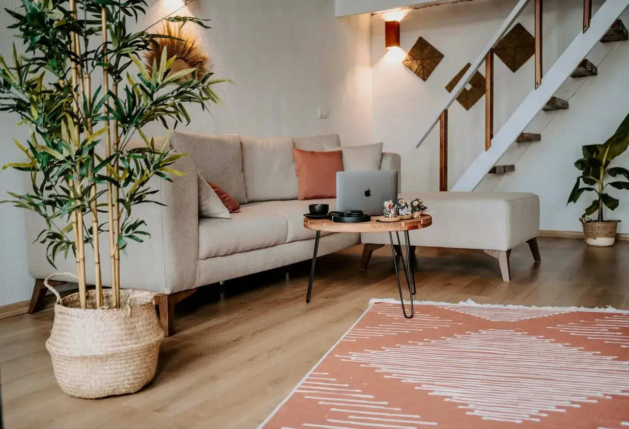 Przytulne, **małe mieszkanie w stylu boho** z beżową kanapą, stolikiem kawowym z laptopem i roślinami. Schody prowadzą na piętro.