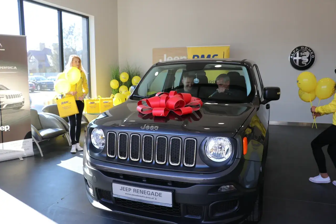 Szczęśliwy zwycięzca odbiera Jeepa Renegade, kiedy final loterii RMF FM. Uroczystość uświetniają żółte balony i czerwona kokarda.