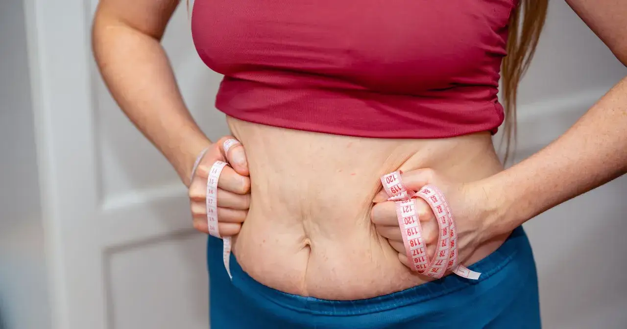 Kobieta ściska brzuch, czując przelewanie w brzuchu. Miarka krawiecka pokazuje obwód.