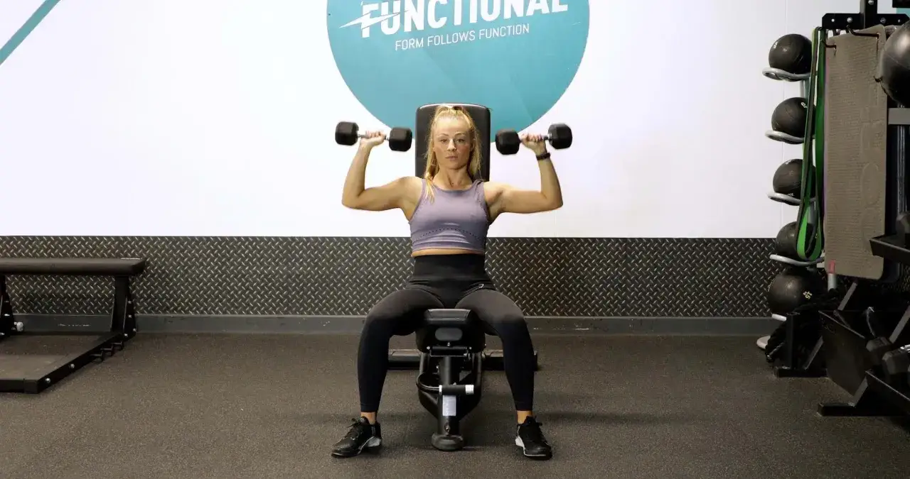 Kobieta wykonuje wyciskanie hantli nad głowę, angażując mięśnie naramienne i triceps.