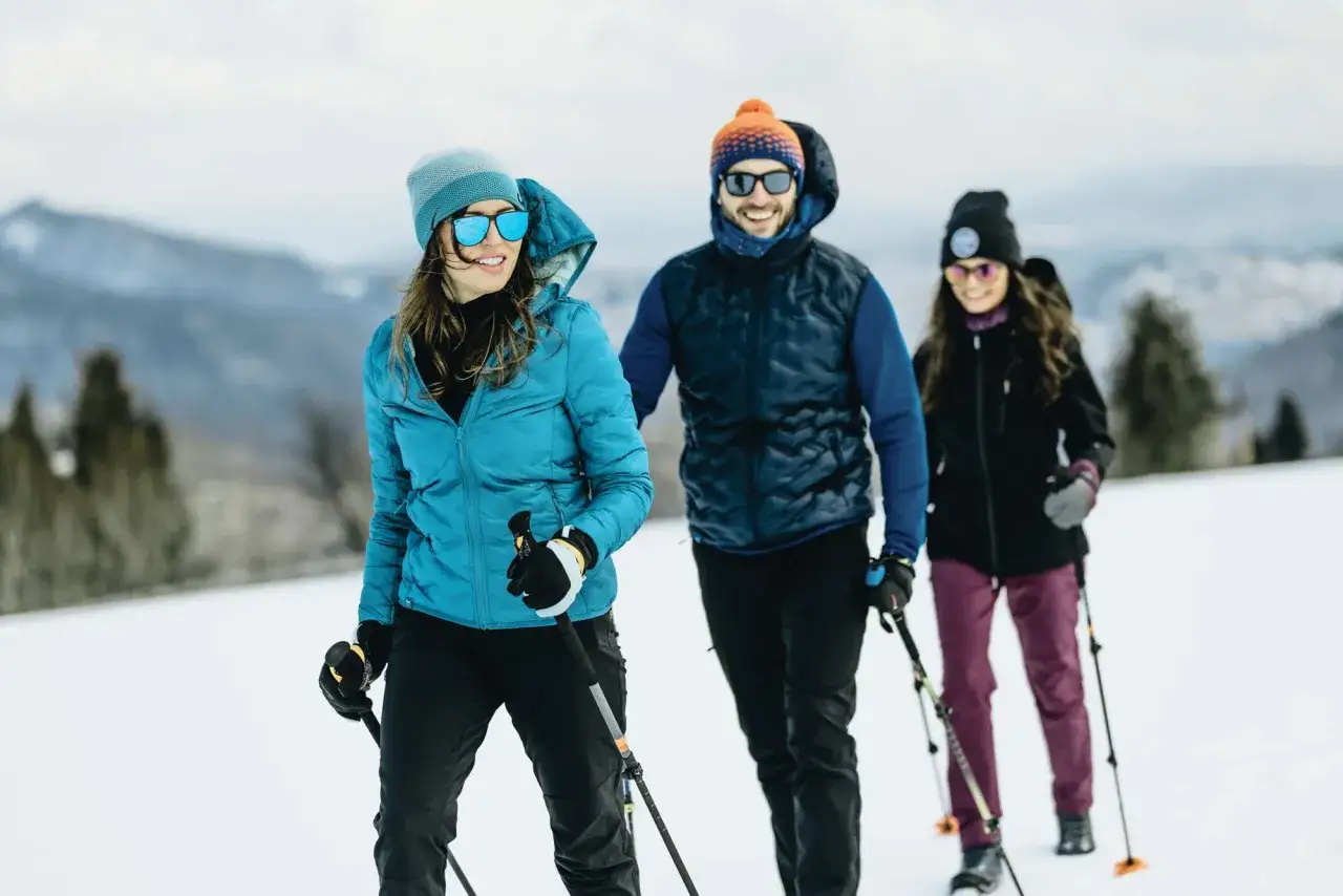 Trzy osoby w zimowych kurtkach i czapkach, z kijkami, pokazują, jak się ubrać w g&oacute;ry zimą.