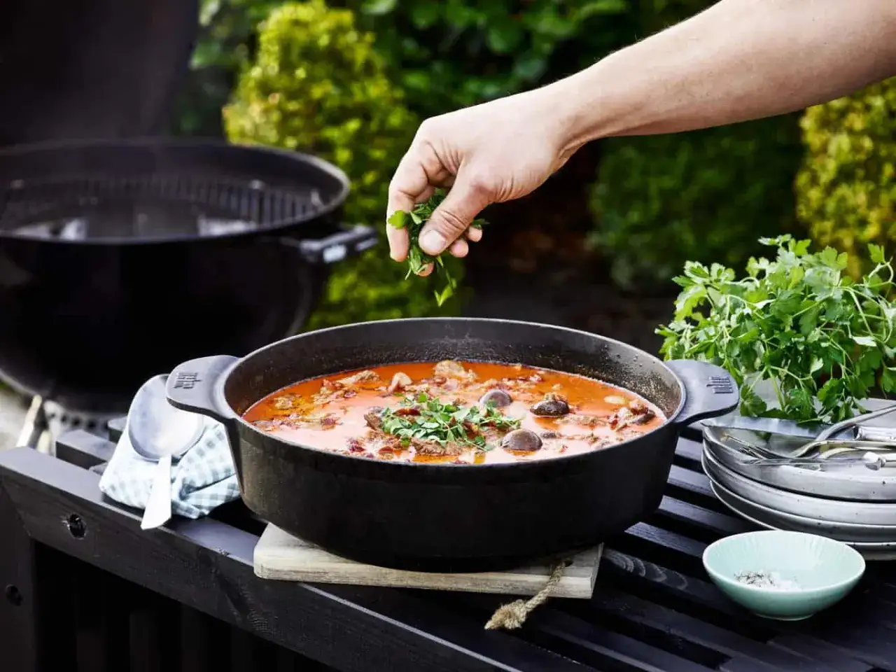 Ręka dodaje zioła do gorącej zupy z grilla w żeliwnym garnku, gotowej do podania na zewnątrz.