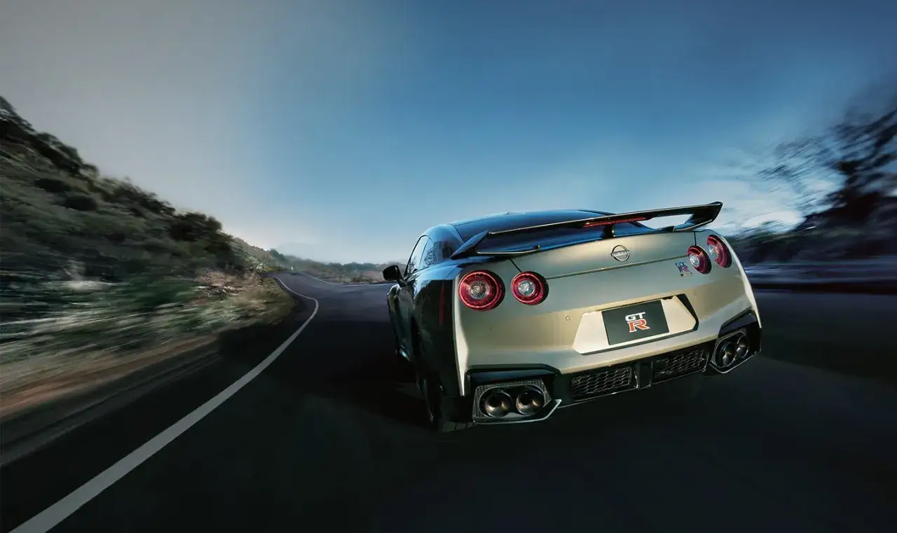Srebrny Nissan GT-R R35 z charakterystycznymi okrągłymi światłami pędzi po krętej drodze wśród gór.