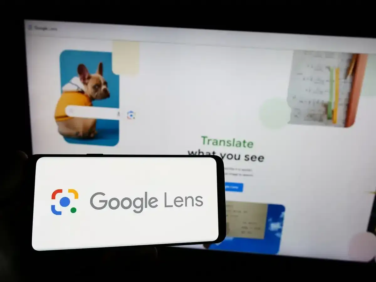 Na ekranie telefonu widnieje logo Google Lens. Za nim, na monitorze, wyświetla się strona z psem w żółtej bluzie i tekstem 
