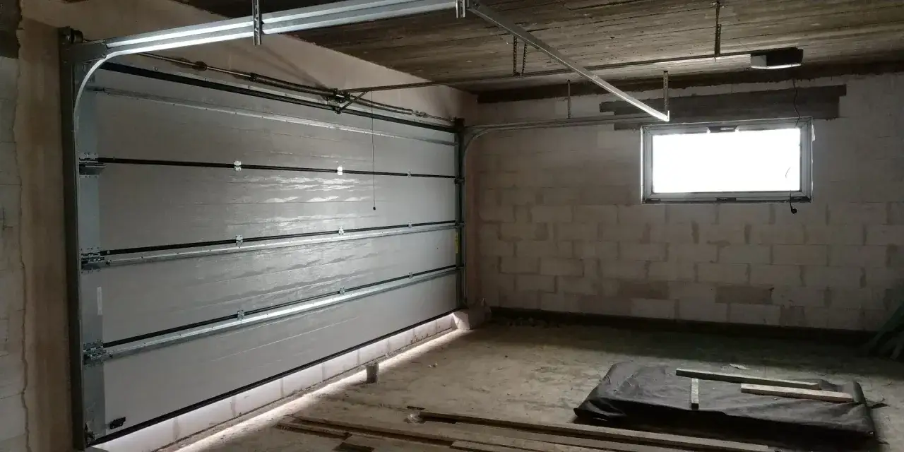 Montaż bramy garażowej w trakcie. Widoczna częściowo otwarta, segmentowa brama, szyny, napęd i okno.
