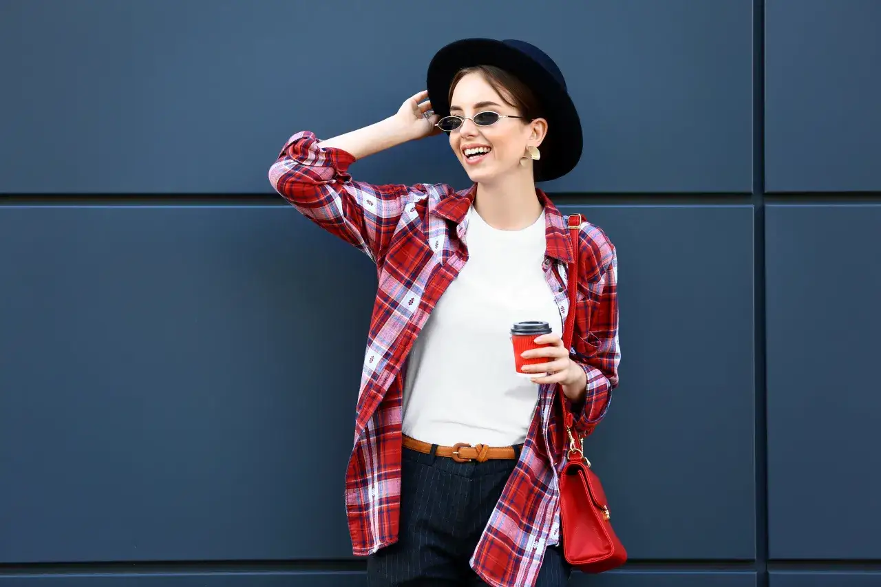Modna dziewczyna w okularach i kapeluszu pokazuje, jak nosić koszulę w kratę damską, tworząc stylowy look z czerwoną torebką.
