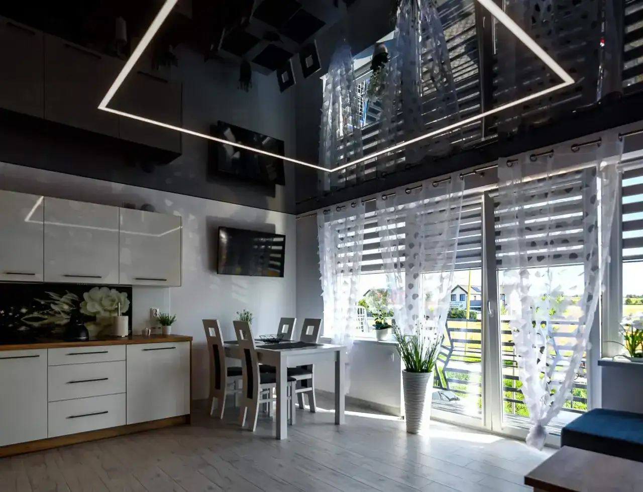 Nowoczesna kuchnia z białymi meblami, stołem i krzesłami. Na czarnym suficie widać subtelne czarne kable, kt&oacute;re dyskretnie wtapiają się w jego fakturę.