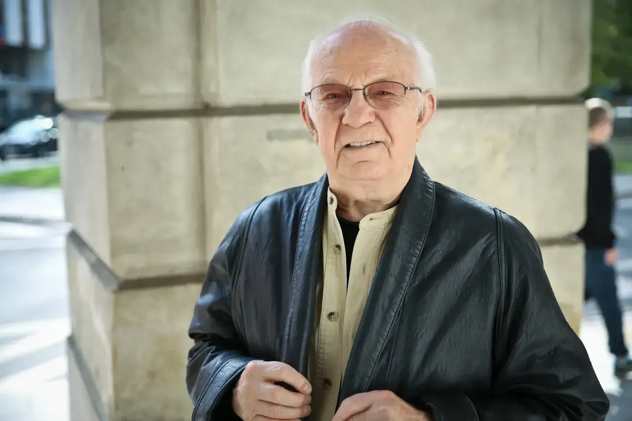 Jerzy Antczak, reżyser znany z niezapomnianych filmów, w czarnej skórzanej kurtce i okularach, pozuje na tle kamiennej architektury.