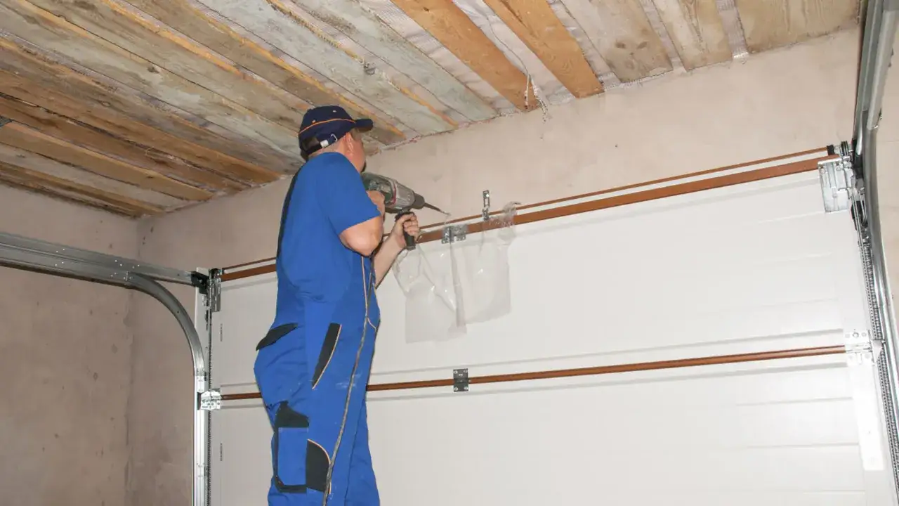 Montaż bramy garażowej w toku. Pracownik w niebieskim kombinezonie wierci otwór w suficie.