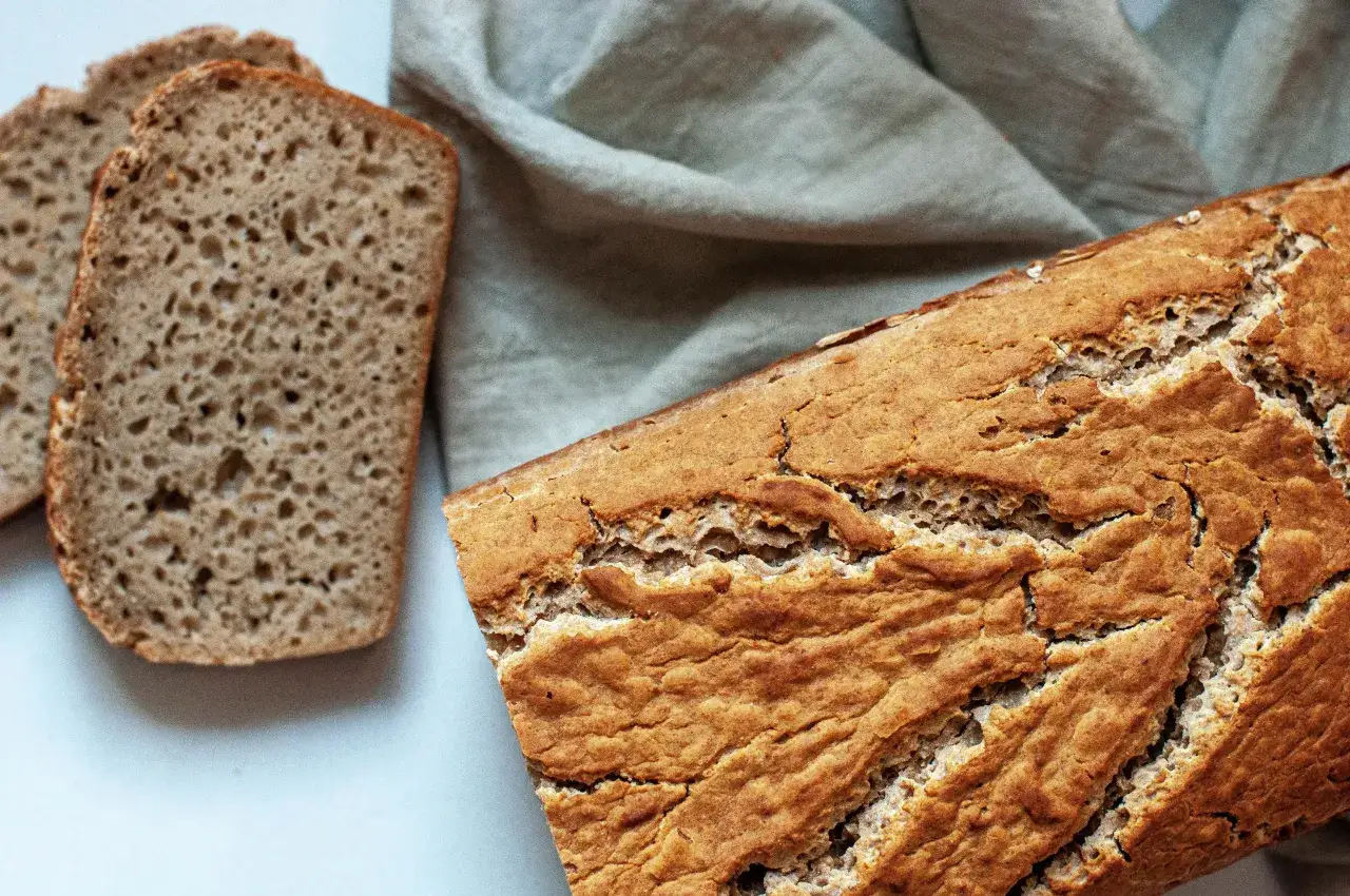 Chleb bezglutenowy przepis: świeżo upieczony bochenek i kromki na jasnym tle, obok lniana ściereczka.