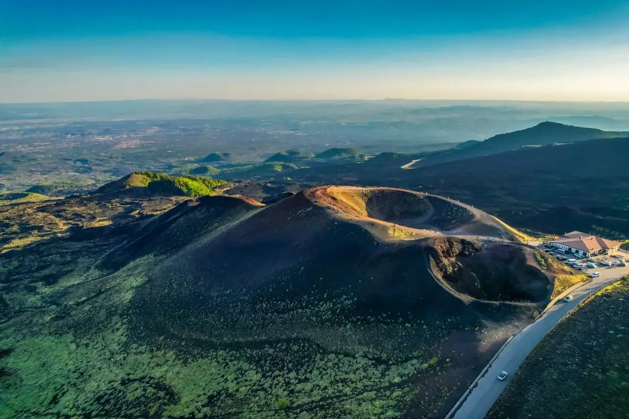 Widok z lotu ptaka na krater wulkanu Etna, gdzie widać kolejkę turyst&oacute;w spacerujących po jego krawędzi. W oddali rozciąga się krajobraz Sycylii.