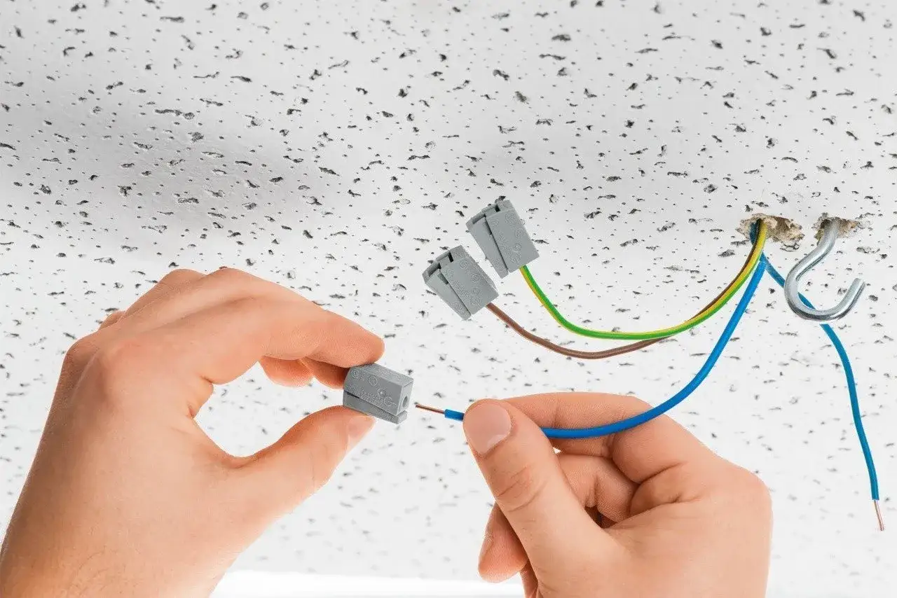 Ręce łączą przewód złączką do lampy sufitowej. Widać instalację elektryczną na suficie podwieszanym.