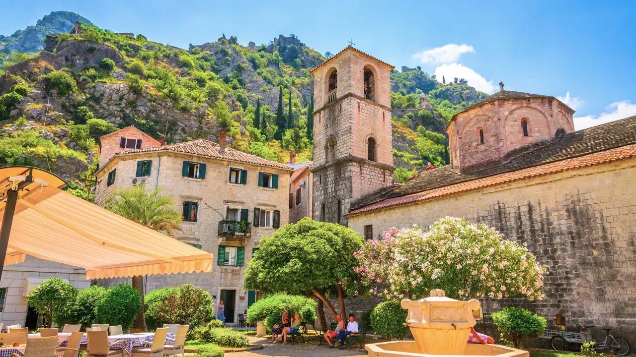 Słoneczna Czarnog&oacute;ra w kwietniu. Urokliwy plac z kamiennymi budynkami, dzwonnicą i kwitnącymi drzewami. Idealna pogoda na relaks.
