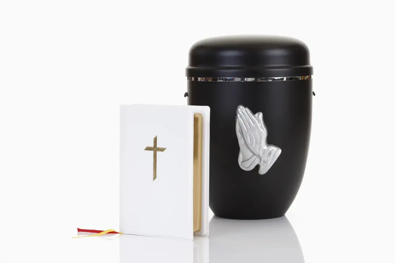 Czarna urna z modlącymi się dłońmi i biała księga z krzyżem. Czy kremacja jest grzechem?