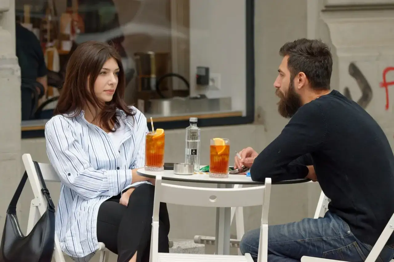 Para siedzi przy stoliku, popijając drinki. Ona uważnie słucha, co m&oacute;wi mężczyzna, kt&oacute;ry pali papierosa.