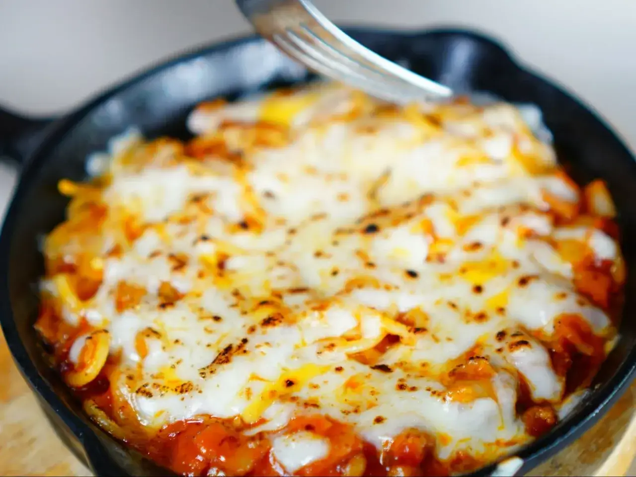 Pyszna zapiekanka makaronowa z ciągnącym się serem, podana w żeliwnej patelni. Widok z g&oacute;ry, widelcem nabierana porcja.