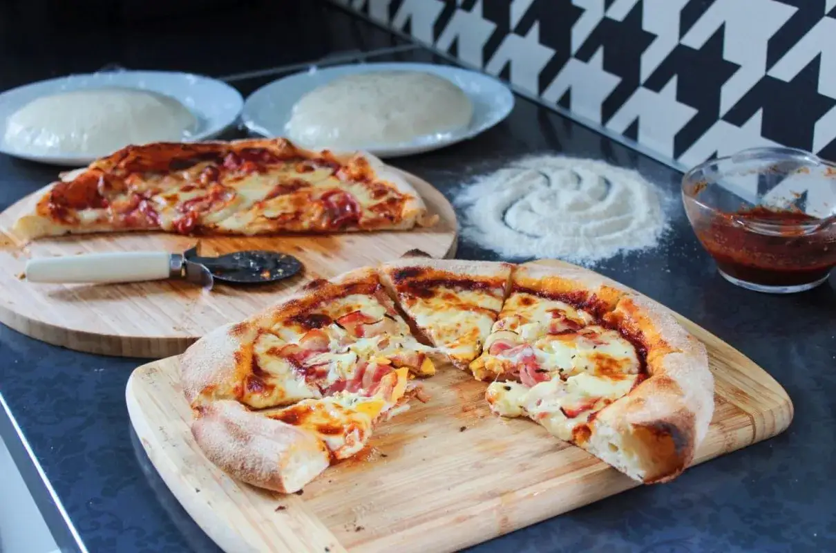 Dwie pyszne pizze na drewnianych deskach, obok mąka i sos. W tle widać kulki ciasta na pizzę, idealne proporcje na domowe wypieki.