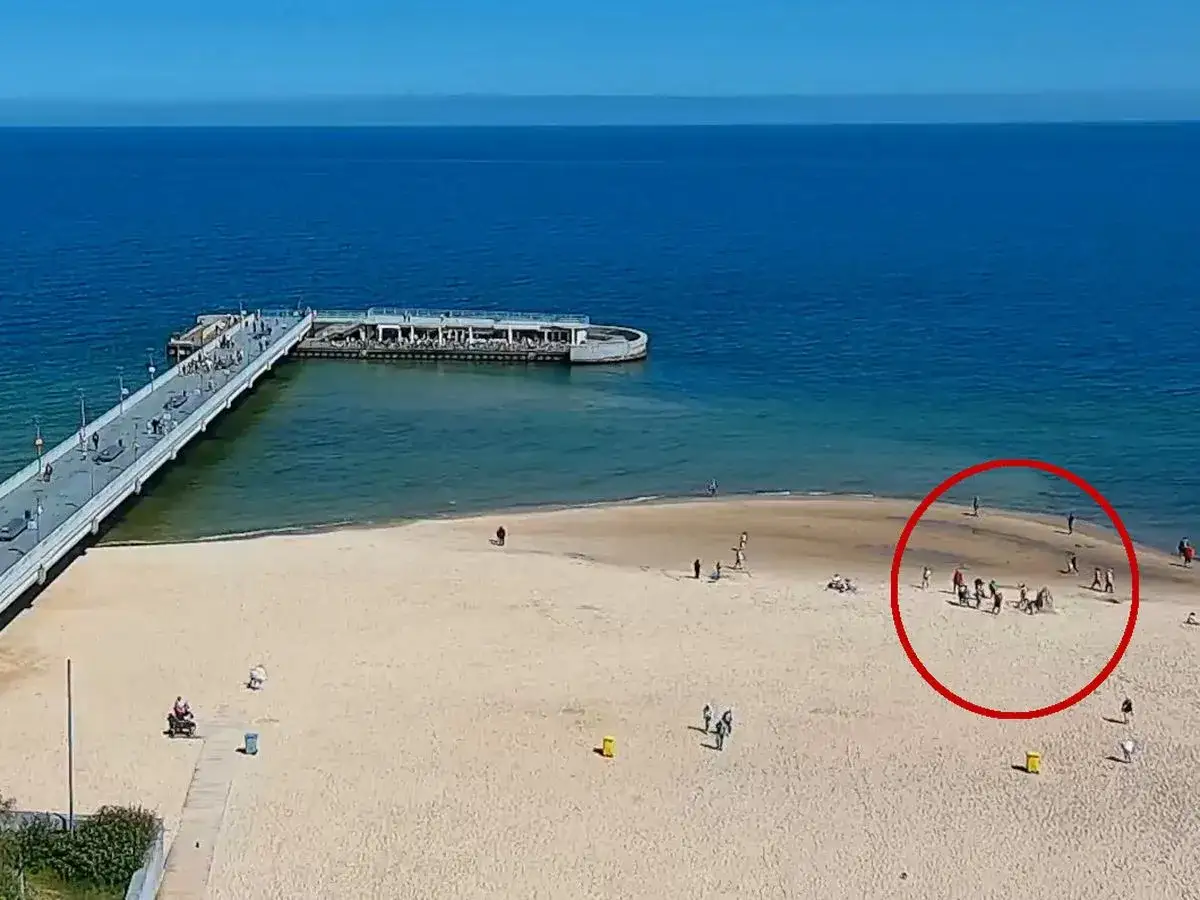 Spacer w Kołobrzegu, z ludźmi na plaży i w wodzie. Czy molo w Kołobrzegu jest płatne? Na razie wygląda na to, że można swobodnie spacerować.