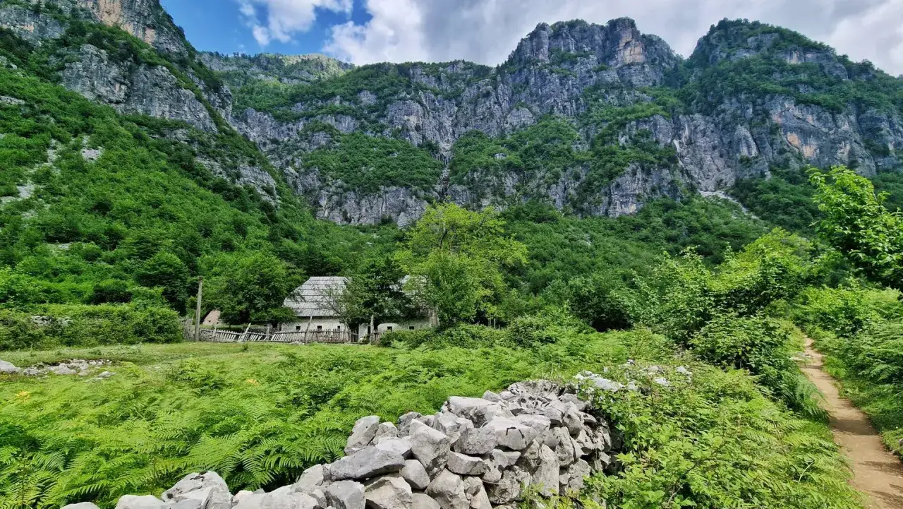 Kamienny murek i ścieżka prowadzą do tradycyjnego domu w Theth, Albania, otoczonego bujną zielenią i majestatycznymi g&oacute;rami.