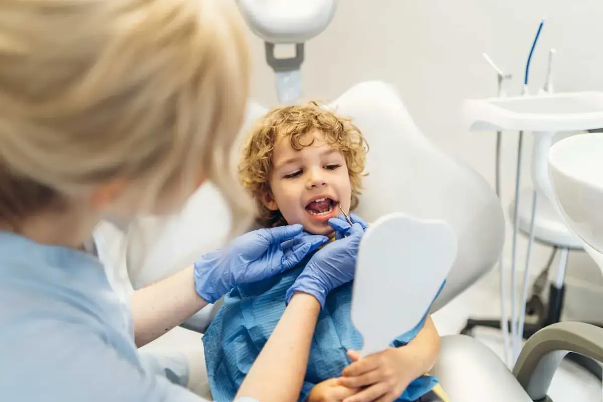 Chłopiec u dentysty, kt&oacute;ry sprawdza, kt&oacute;re zęby są mleczne. Dziecko patrzy w lusterko, a dentystka bada jego uśmiech.