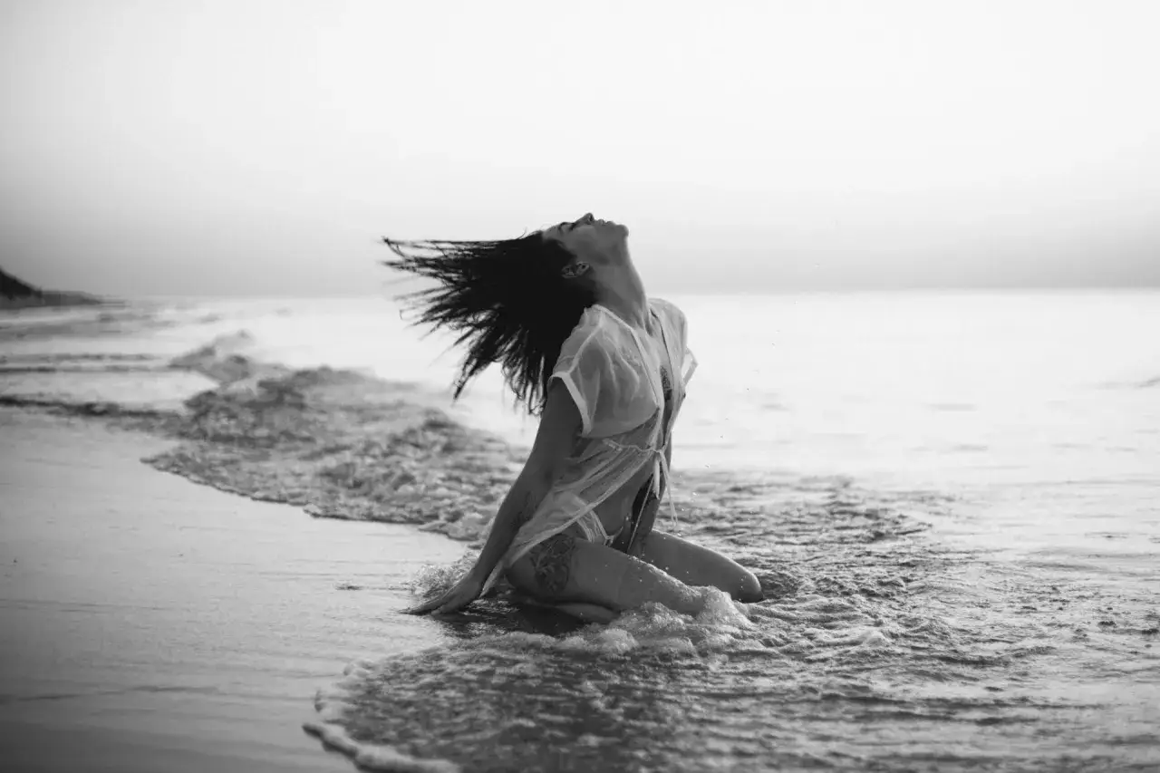 Kobieta w zwiewnej sukience wykonuje fajne pozy do zdjęć nad morzem, włosy rozwiane przez wiatr, fale obmywają jej nogi.