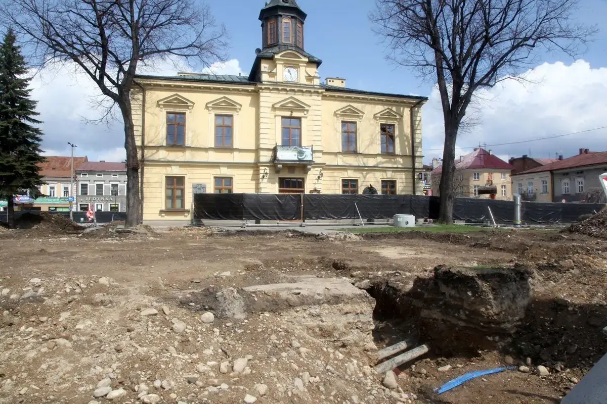 Plac budowy w Nowym Targu. W tle żółty budynek ratusza, drzewa i inne kamienice. Trwają prace ziemne, widoczne wykopy i rury.