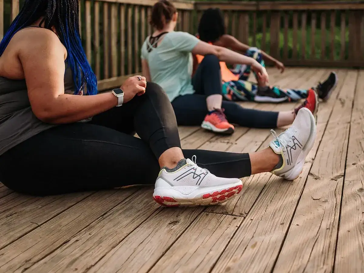Trzy kobiety w strojach sportowych siedzą na drewnianym tarasie, przygotowując się do treningu. To inspiracja, jak nauczyć się biegać.