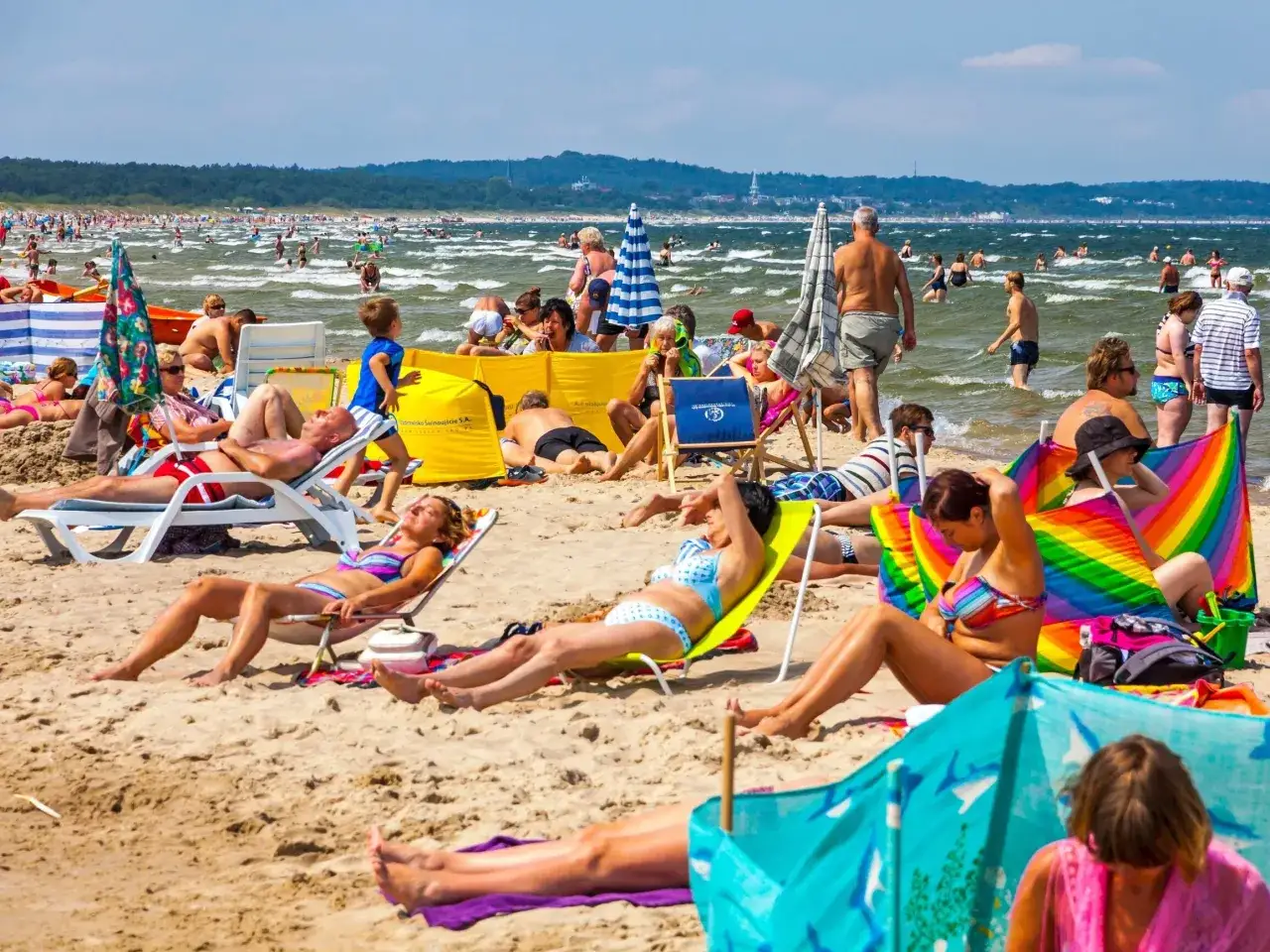 Tłum ludzi na plaży, opalanie się i kąpiel w morzu. Szukasz miejsca, gdzie nad morzem jest najtaniej? Ta plaża wygląda na popularną i pełną życia.