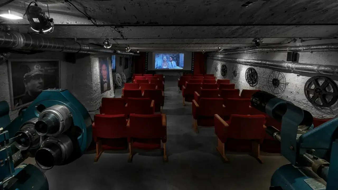 Kameralna sala kinowa z czerwonymi fotelami i zabytkowym projektorem. W tle ekran z filmem i dekoracje nawiązujące do historii kina, jakby to było pierwsze kino w Polsce.