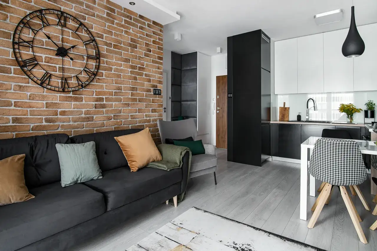 Nowoczesna ozdobna ściana do salonu z cegły, z dużym zegarem, obok czarnej kanapy i szarego fotela.