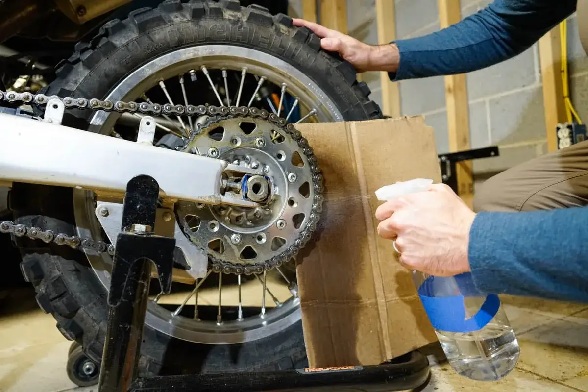 Osoba wykonuje smarowanie łańcucha motocyklowego, rozpylając płyn na zębatkę i łańcuch.