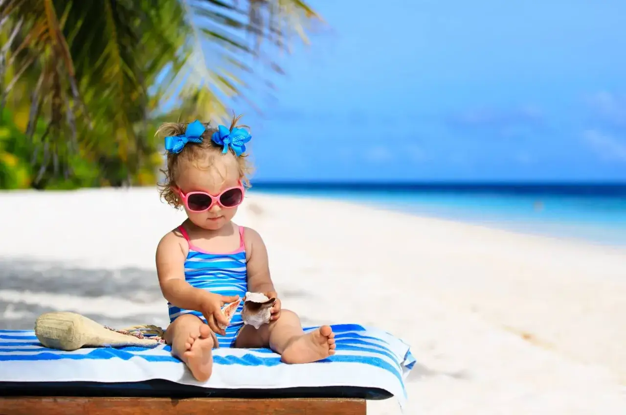 Mała dziewczynka w okularach przeciwsłonecznych i kostiumie kąpielowym siedzi na plaży, bawiąc się muszlami. Idealne miejsce, gdzie na wakacje z niemowlakiem.