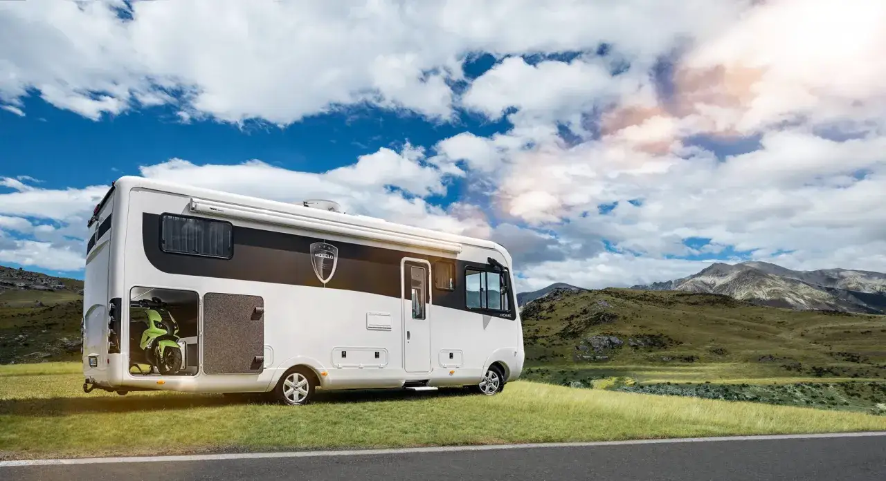 Biały kamper z zielonym skuterem w garażu, zaparkowany na trawie z widokiem na g&oacute;ry. Imponująca wysokość kampera pozwala na wygodne przechowywanie.